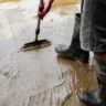 Osoba w gumowcach czyści szczotką zabłoconą, mokrą betonową posadzkę